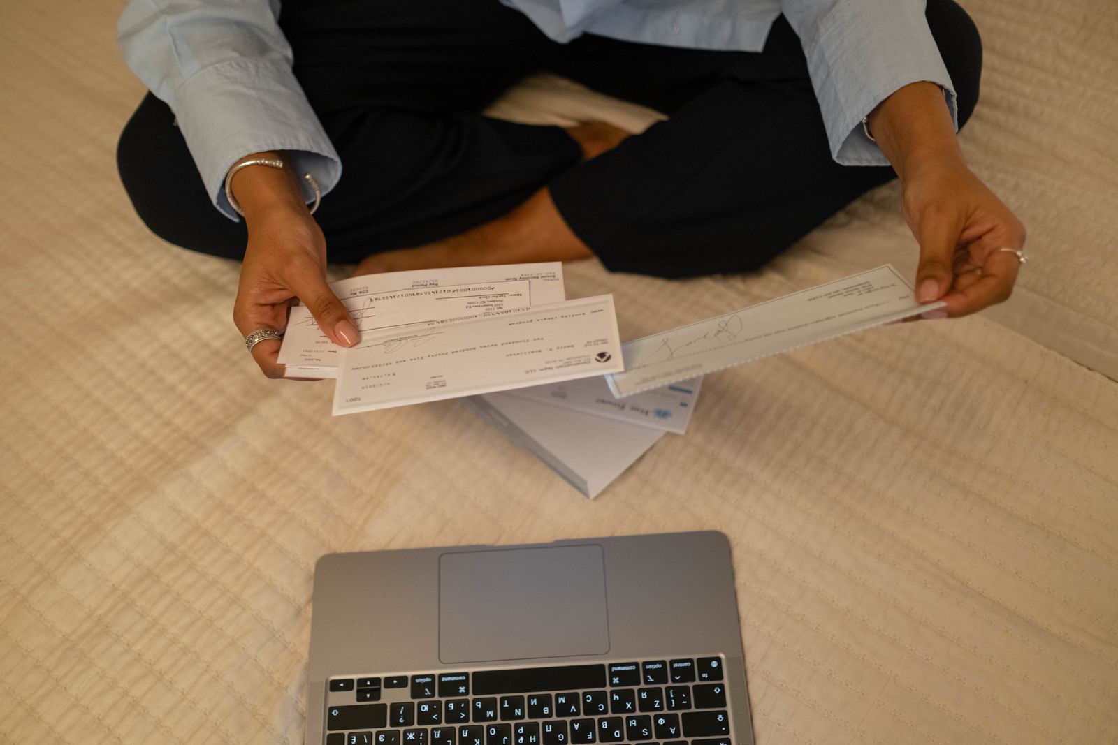 A person sitting cross-legged on a bed, holding paper checks and a pay stub above an open laptop