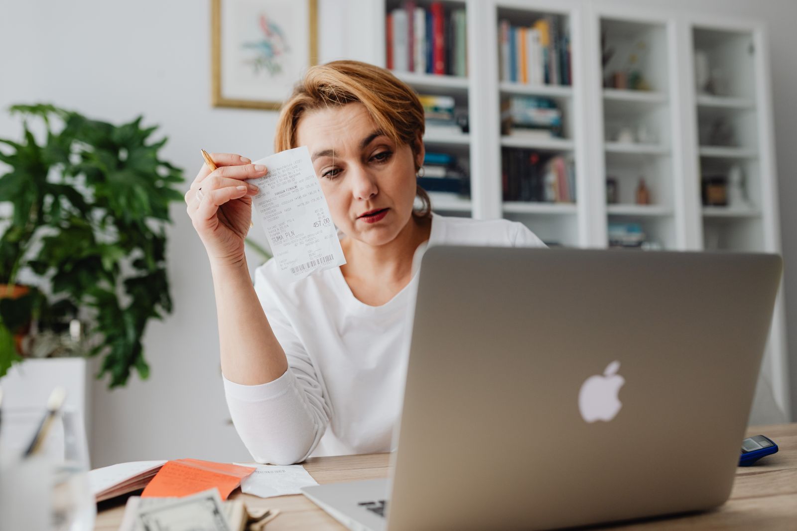 A woman at a laptop holding up a store receipt, with more receipts and dollar bills on the desk