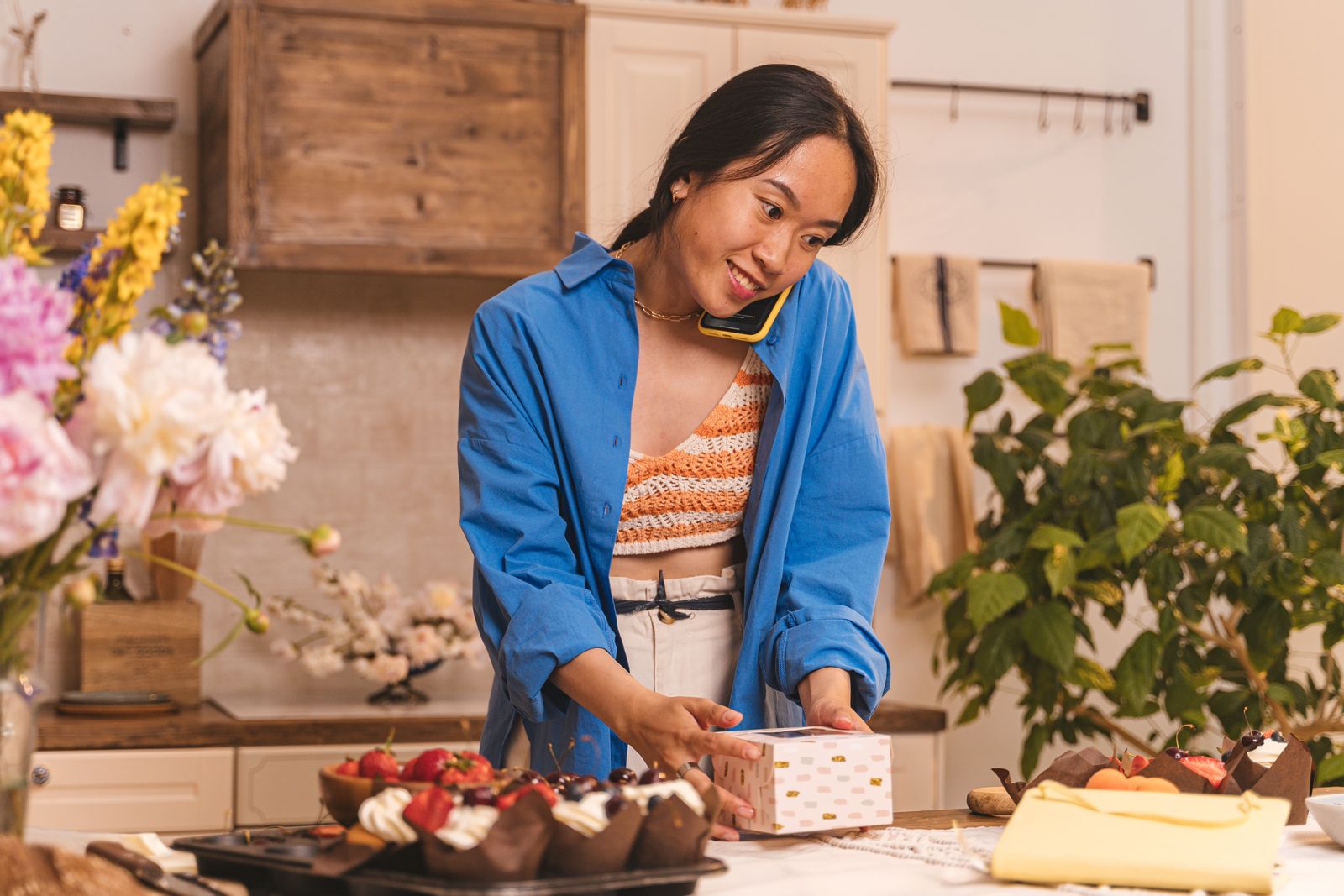 A woman on a phone call packing a pastry box at a counter with muffins and fresh fruit in a small bakery kitchen