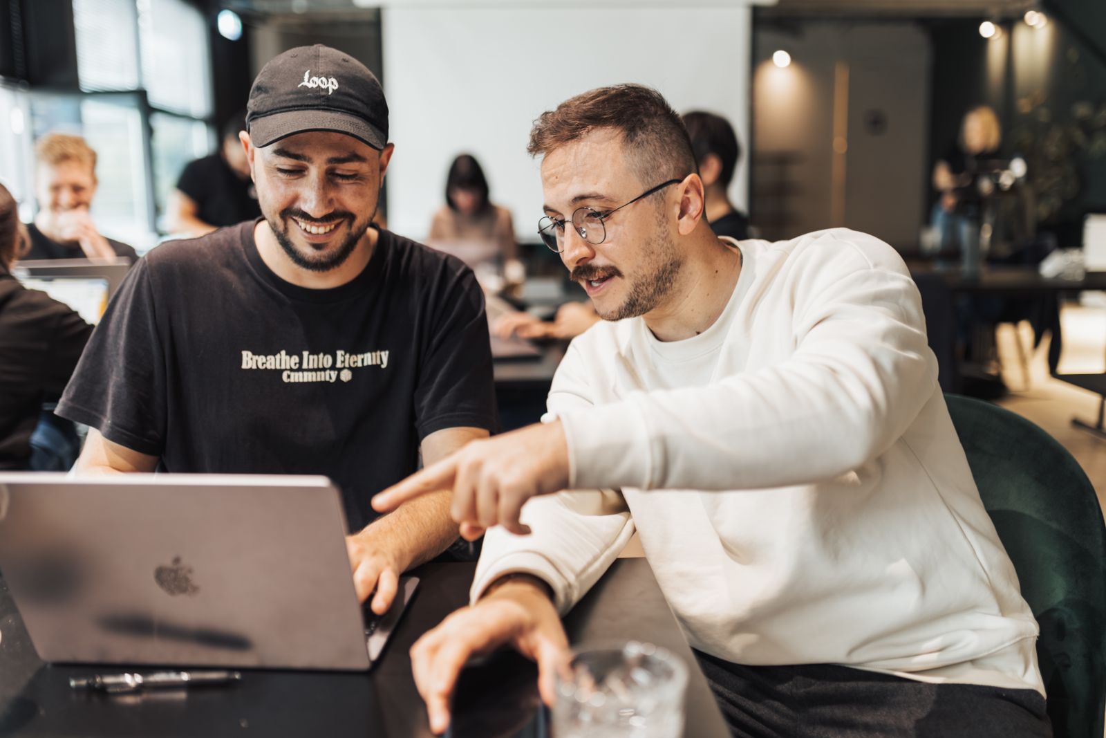 Two young men at a shared table in a busy office, one pointing at the other's laptop screen