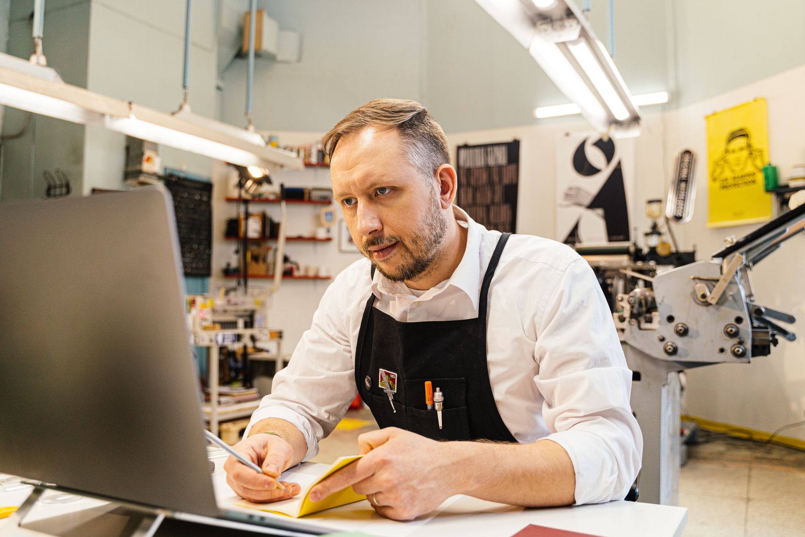 A man in a work apron taking notes while looking at a laptop in a print shop, with a printing press behind him