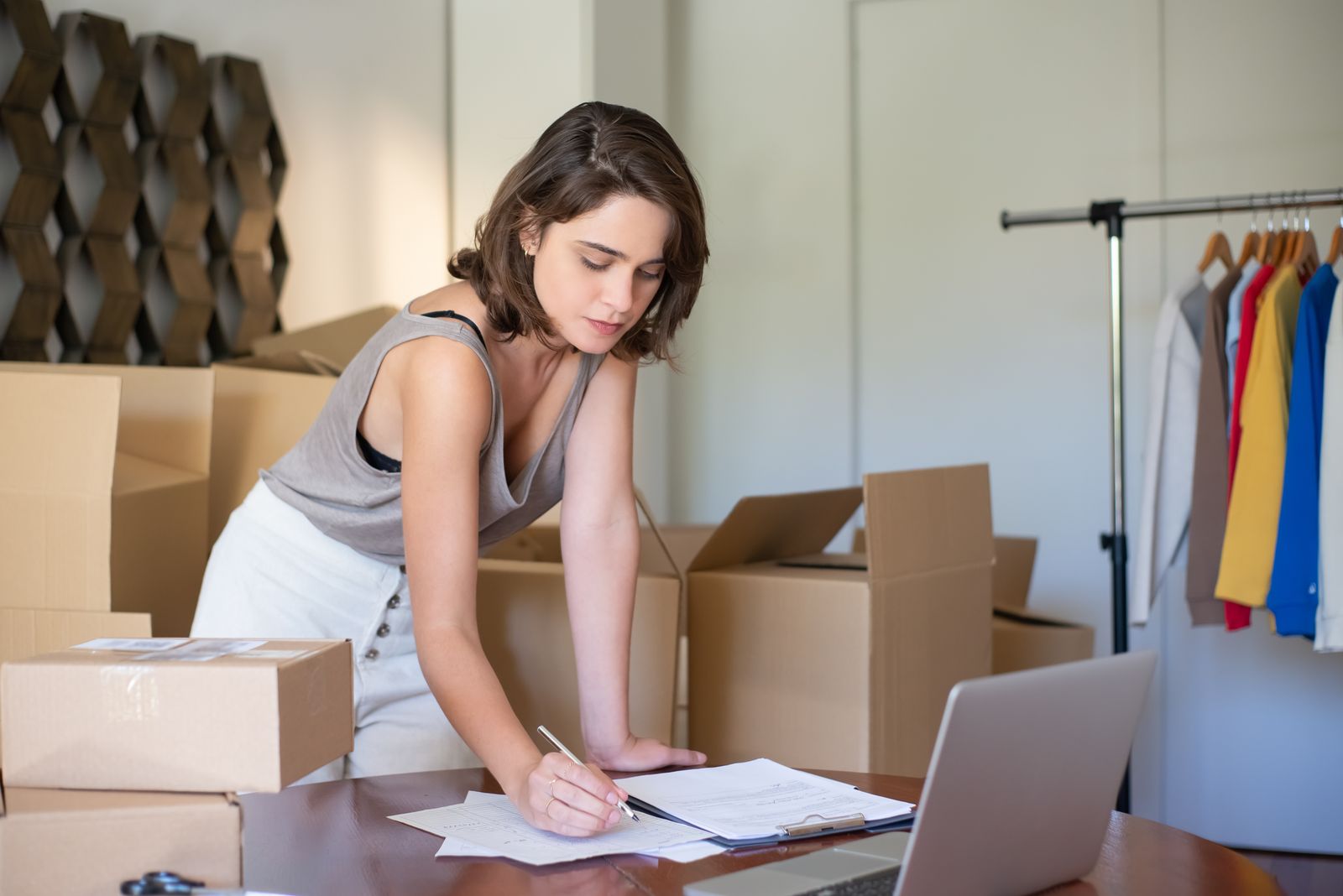 A woman leaning over a table to fill in paperwork beside a laptop, with shipping boxes and a rack of shirts behind her