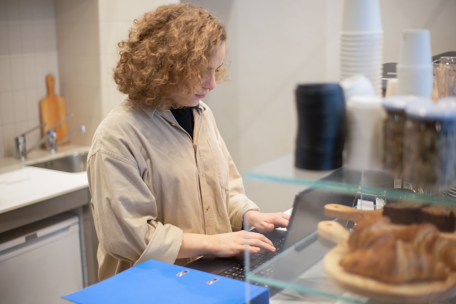 A woman typing on a laptop behind a café counter, with pastries on a glass shelf in the foreground