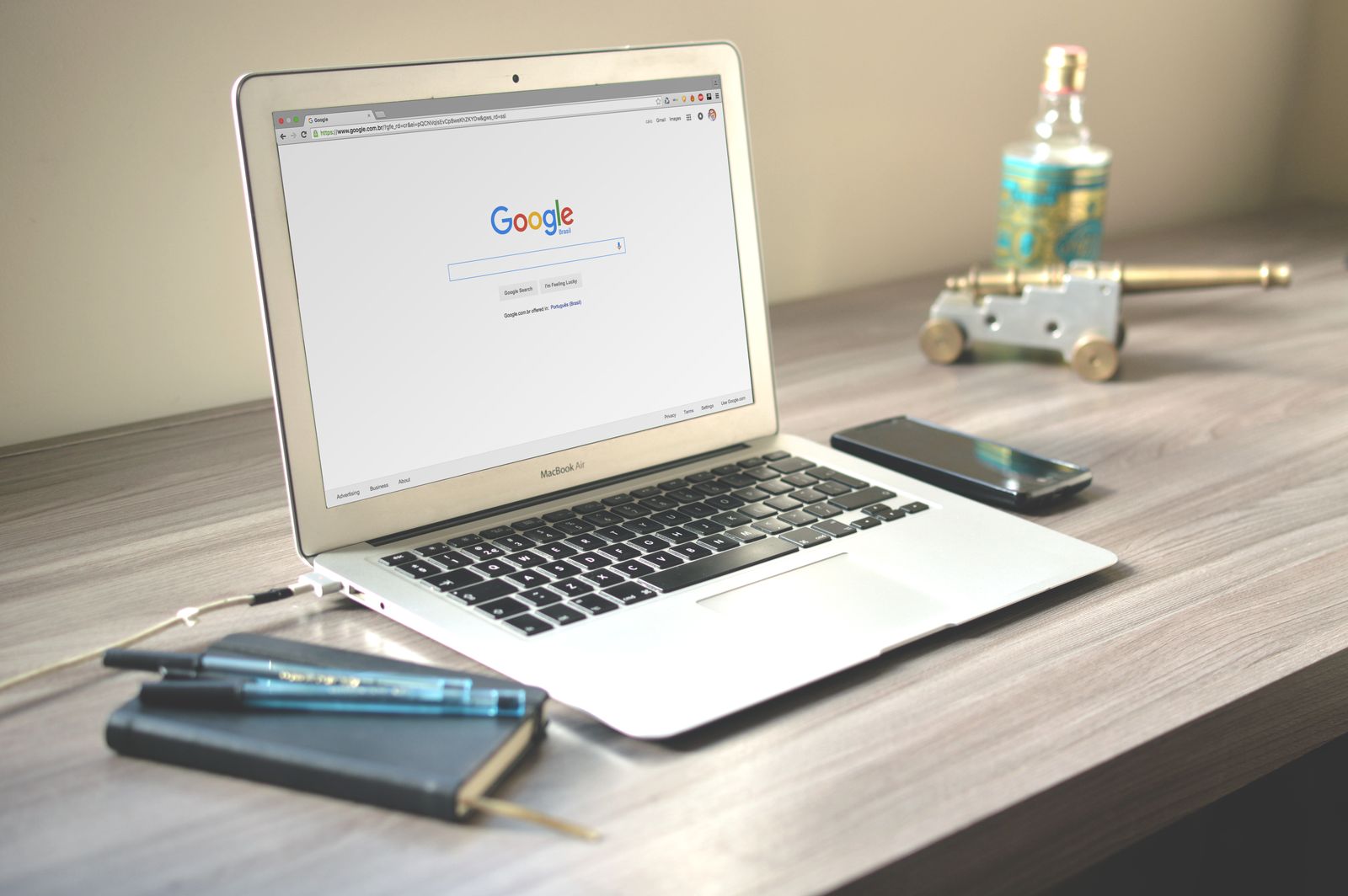 A laptop on a wooden desk open to the Google search page, next to a smartphone and a notebook with two pens