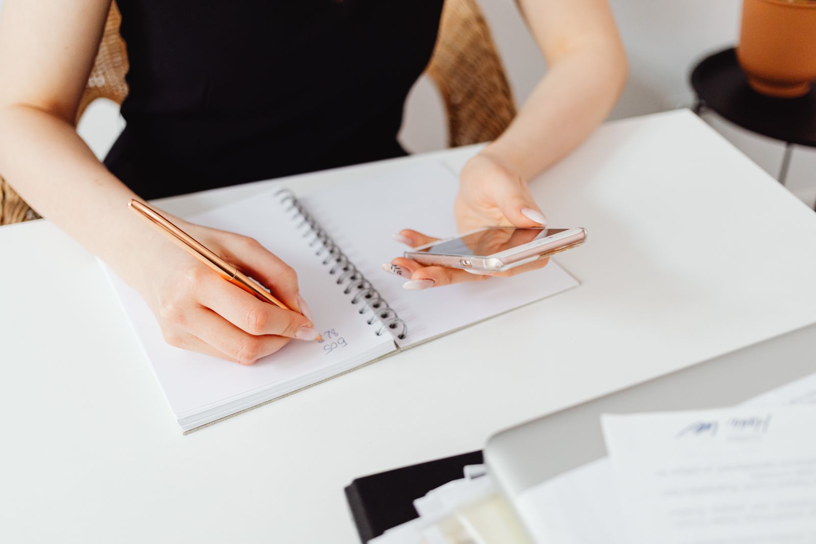 A woman planning in a notebook while holding a smartphone at a white desk