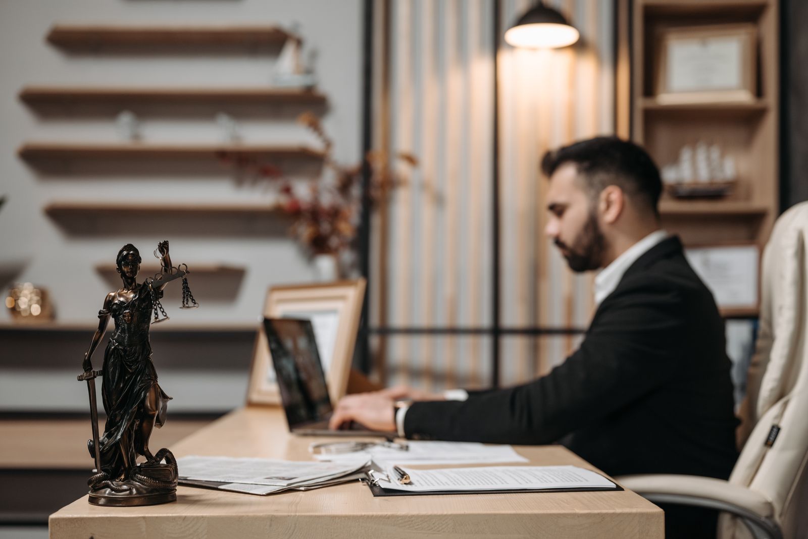 An attorney typing on a laptop at a wooden desk with papers, a Lady Justice statuette in the foreground