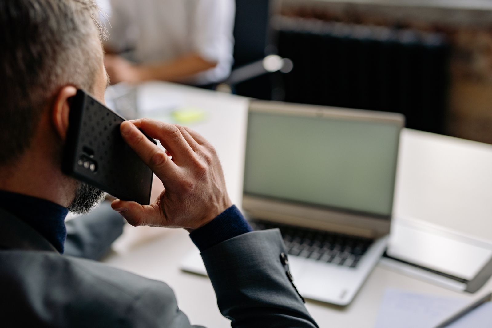 A man in a suit on a phone call at a desk with an open laptop