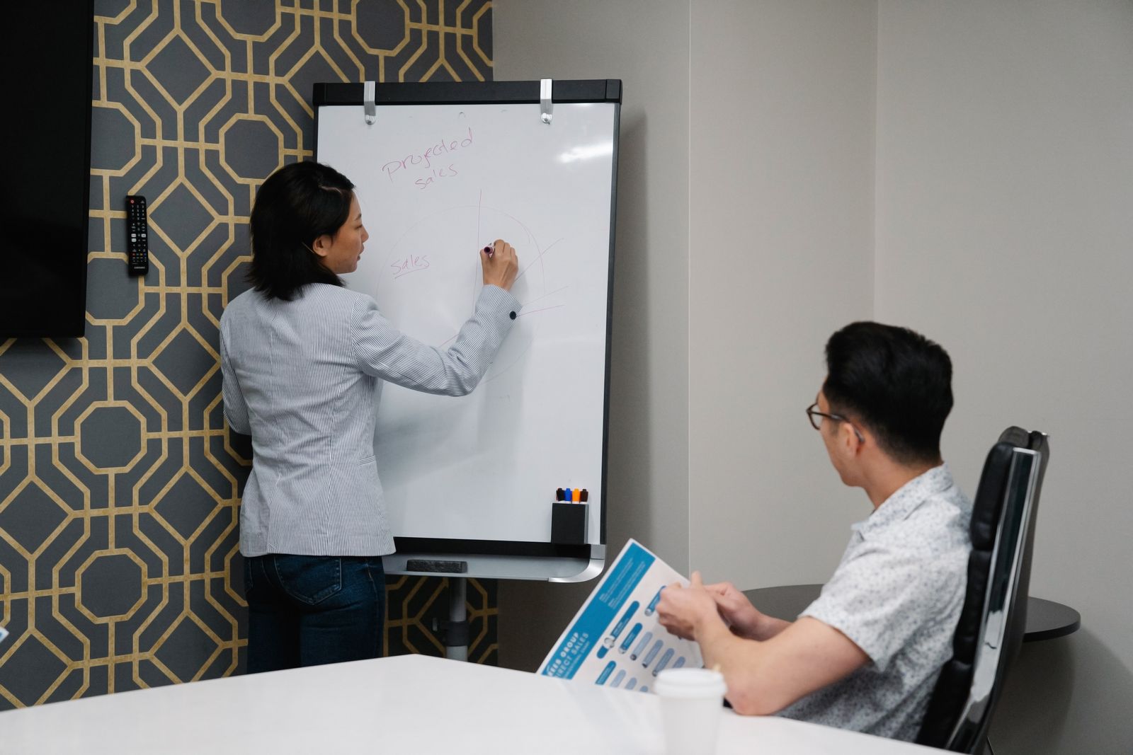 A woman drawing a sales chart on a whiteboard while a colleague holding a report watches