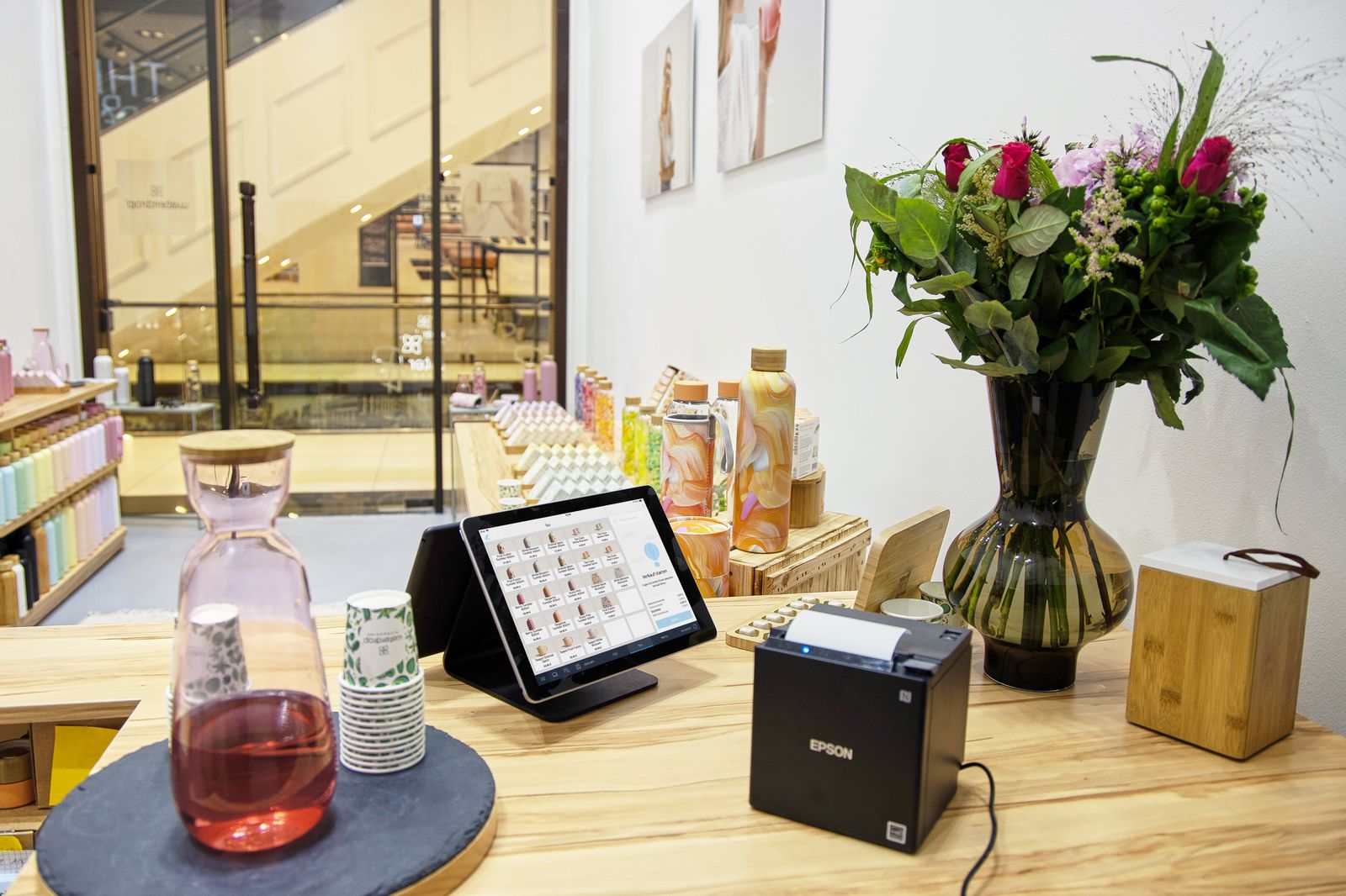 A tablet point-of-sale screen on a stand and a black Epson receipt printer on a light wooden shop counter, with a vase of flowers and shelves of bottles behind
