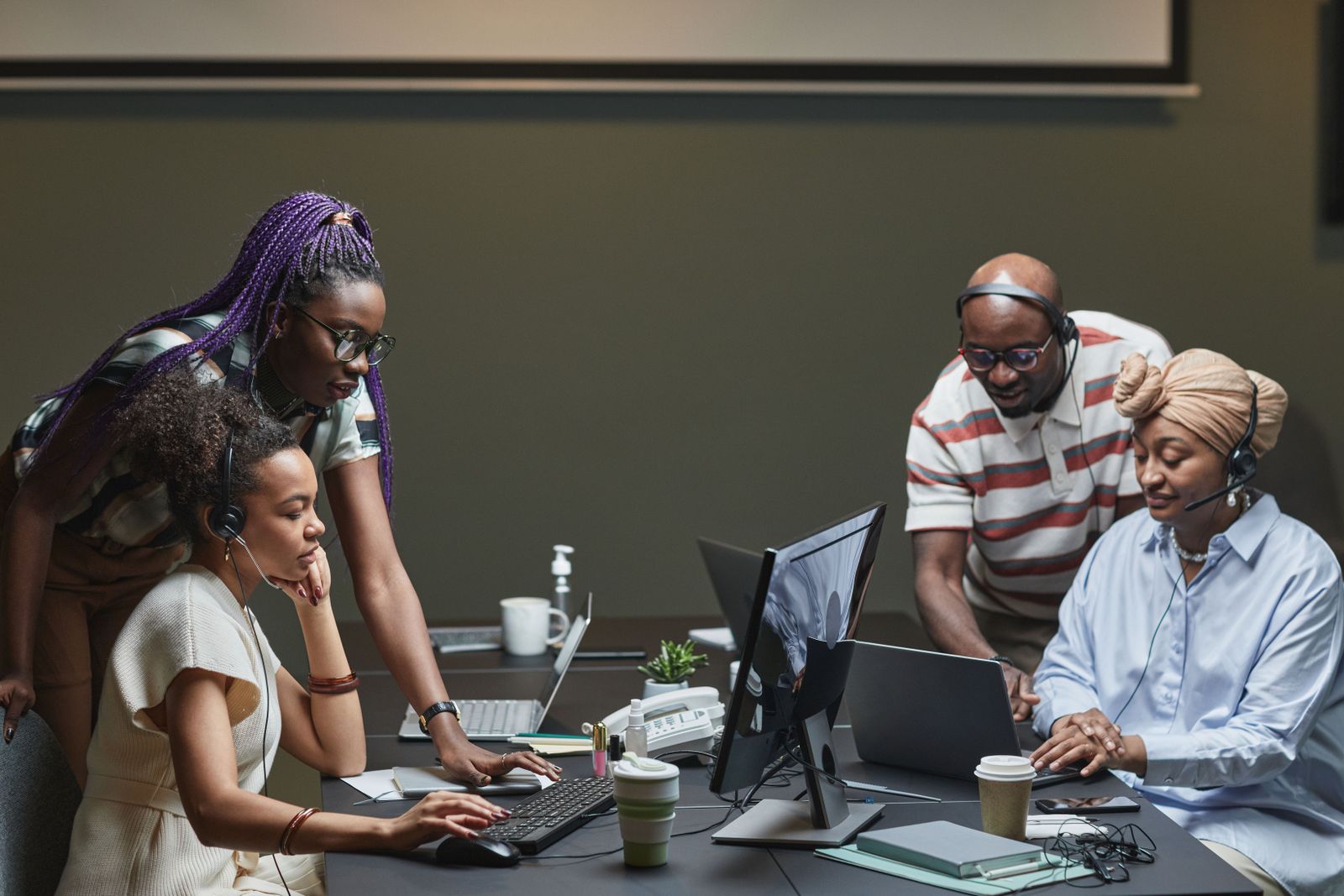 Four colleagues, three wearing headsets, working together at a shared desk with computers and a desk phone