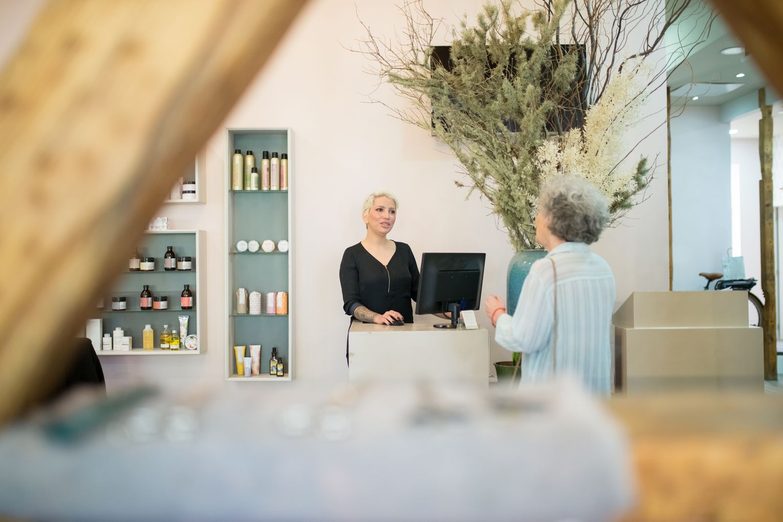 A salon receptionist at a computer at the front desk talking with a gray-haired client, with shelves of hair products behind her