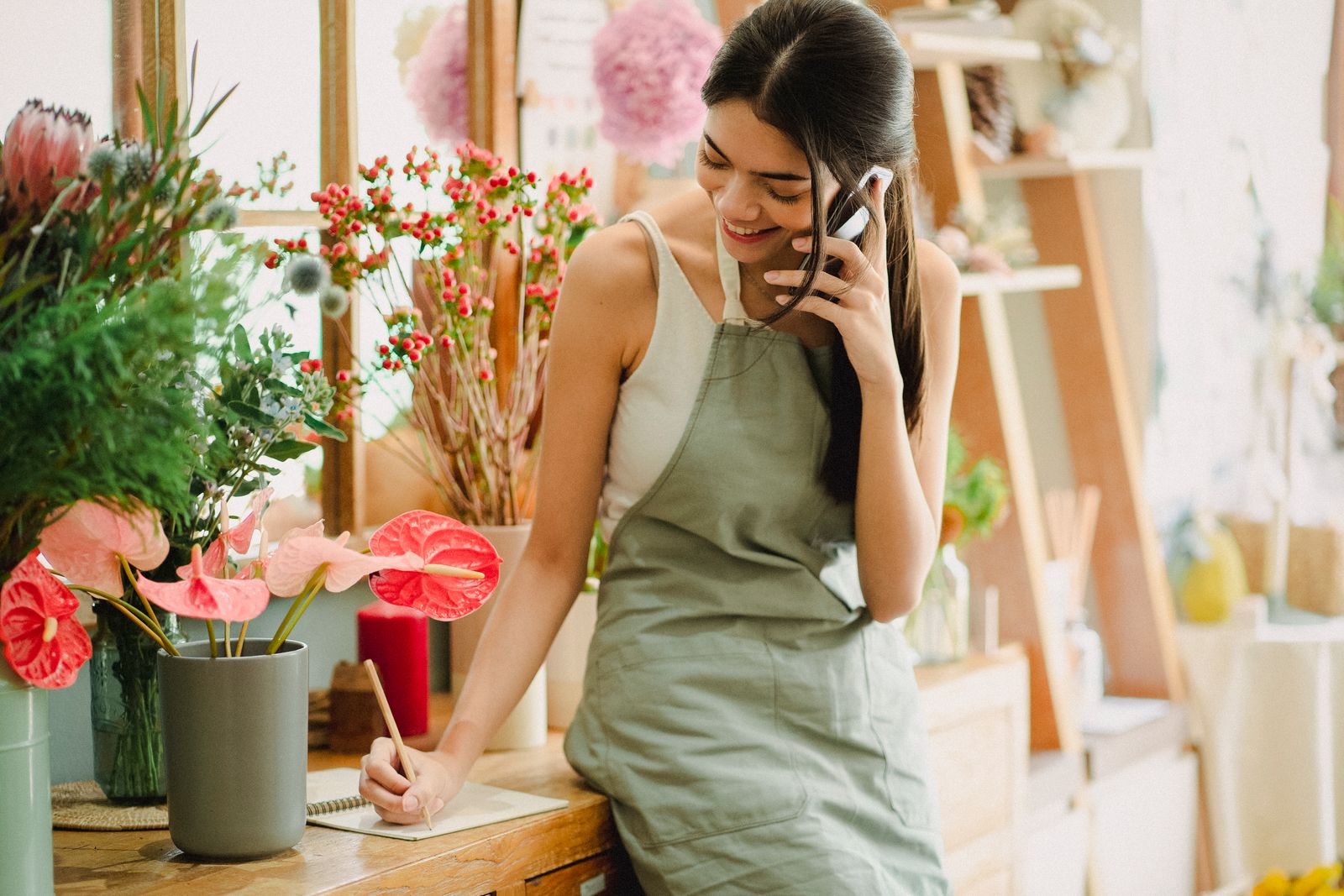 A florist in a green apron smiling as she takes a call on her mobile phone and writes a note at the shop counter