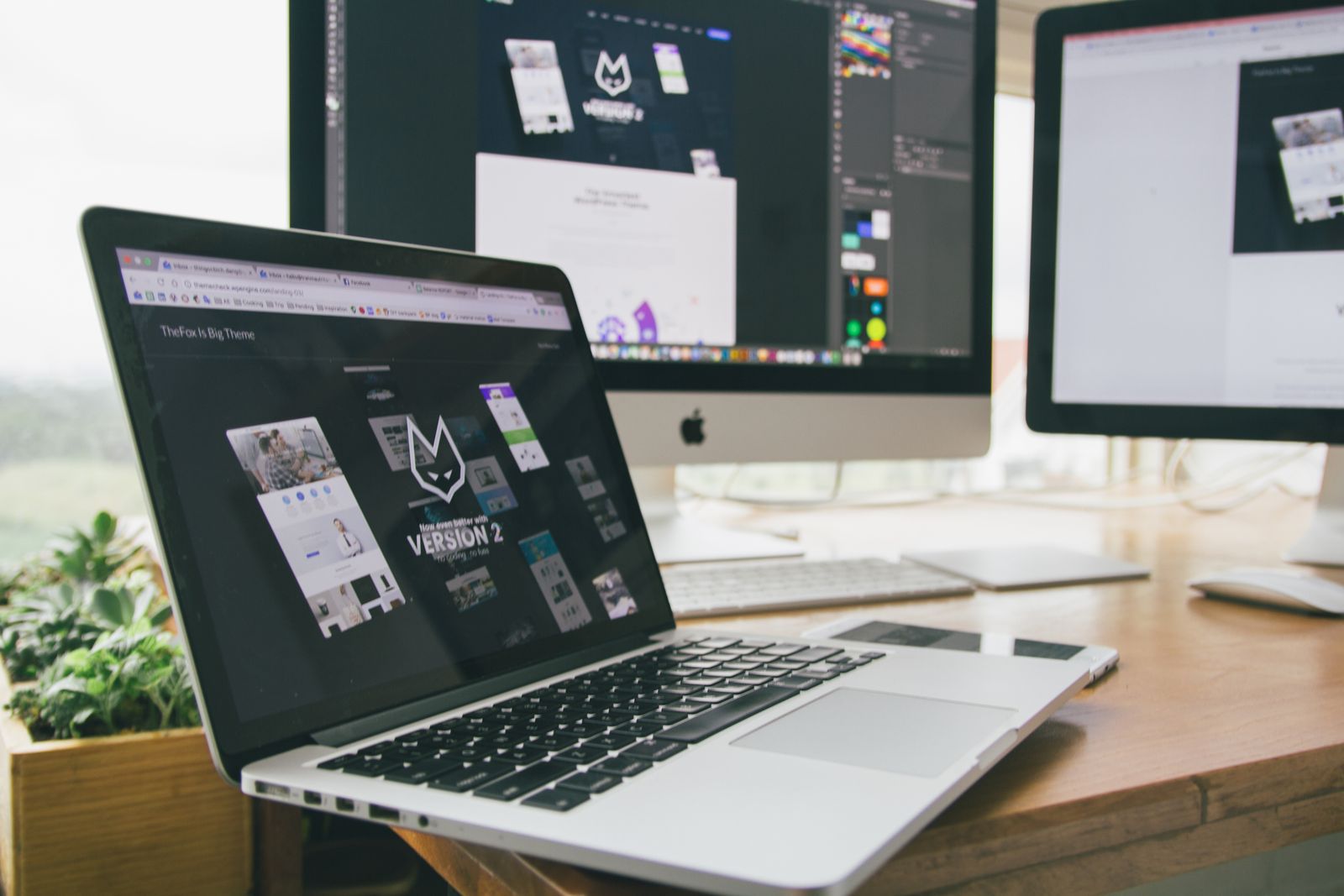 A laptop showing a website theme's landing page on a desk, with two monitors behind it open in a design app